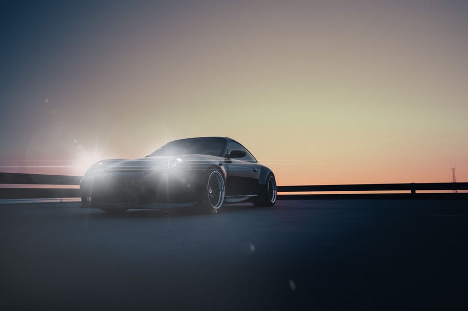 Dark sports coupe on an open highway at dusk, headlights flaring against a fading orange sky