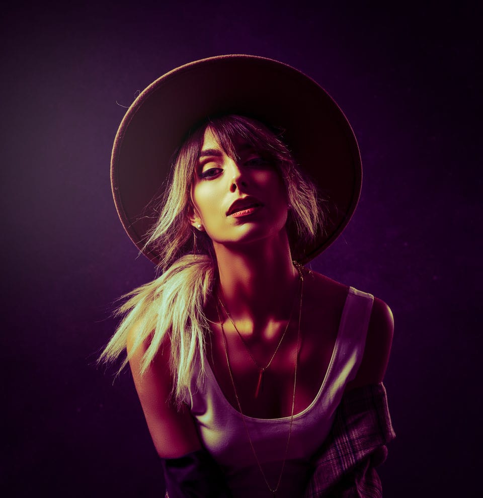 Portrait of a woman in a wide-brimmed hat, lit in violet and warm gold