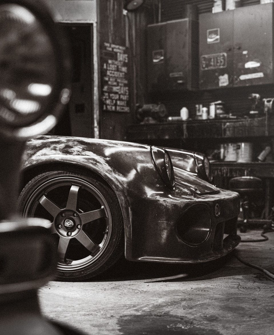 Raw metal car body on deep-dish wheels inside a dim, cluttered garage, in black and white